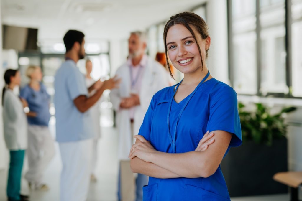 a young nurse in blue scrubs smiling and standing in bright hallway with other medical professionals in background