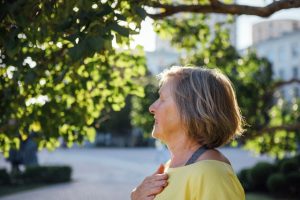 Elderly woman with gray hair in a yellow shirt stands in a park, with hand placed on chest
