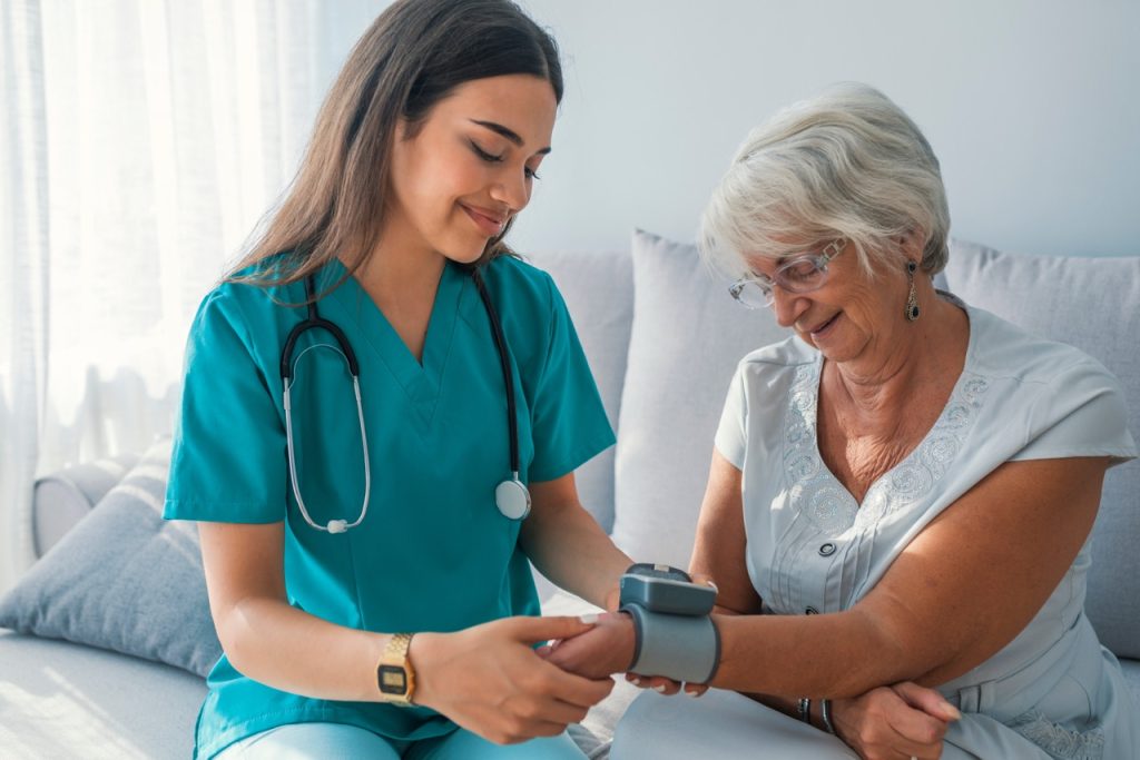 young woman CNA taking blood pressure of senior woman sitting on couch in home